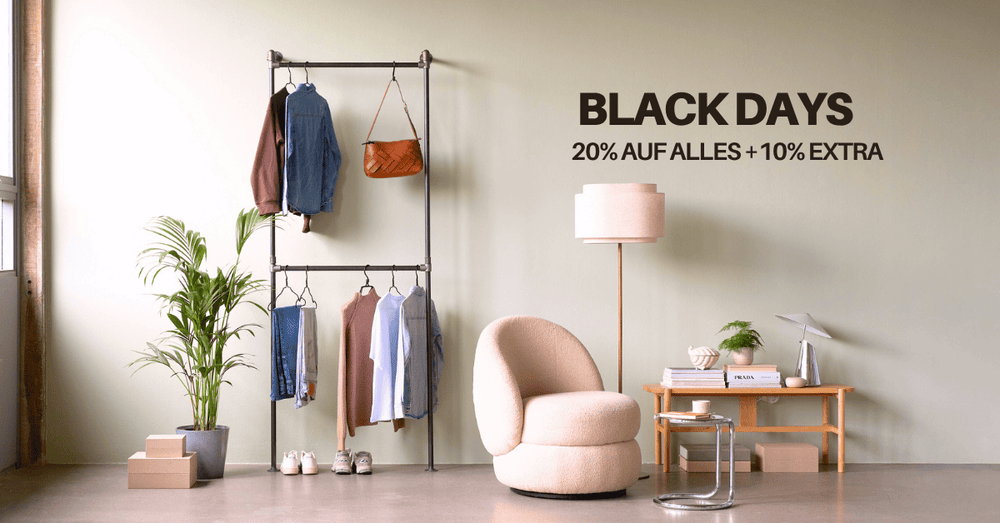 Heller Raum mit Kleidung auf Stahlstangen, grünem Pflanzenpot, beiger Lounge-Kugelcouch und Holzschreibtisch mit Lampe an heller Wand