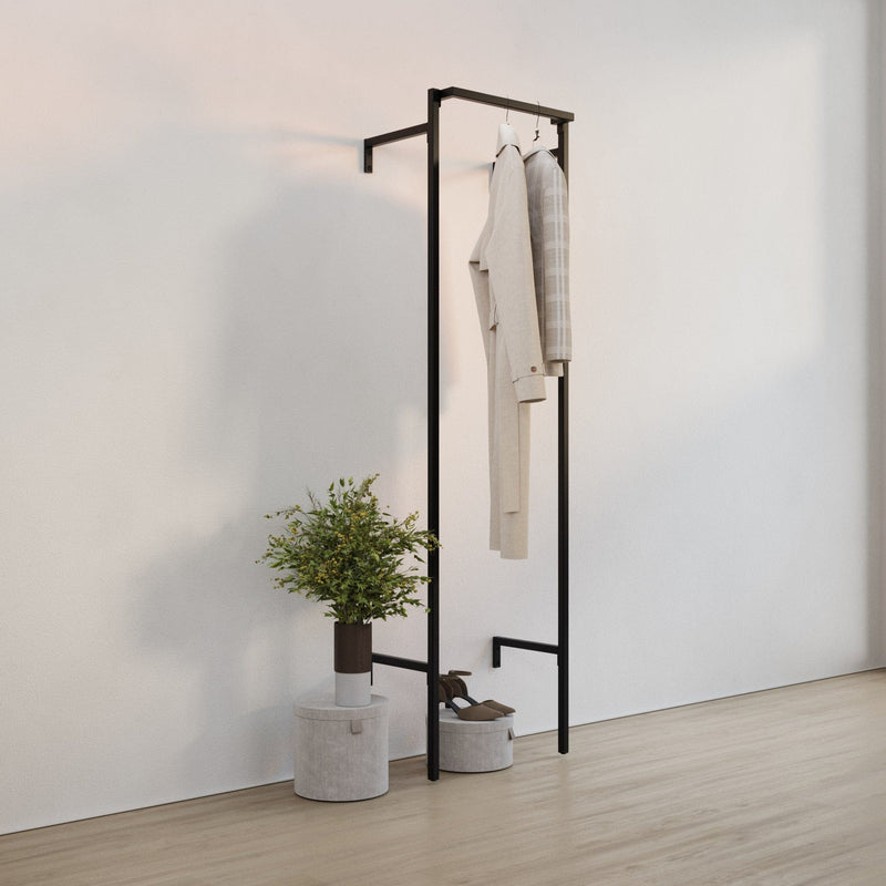 Schlichter schwarzer Garderobenständer an weißer Wand mit zwei Jacken, grauer Sitzbank und Pflanze auf hellem Boden