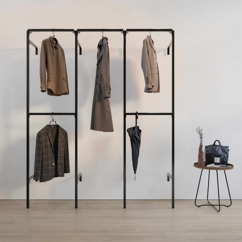 Schwarzes Kleidergestell mit verschiedenen Manteln, Jacken, einem Regenschirm und daneben kleiner Tisch mit Tasche und Deko vor hellem Hintergrund