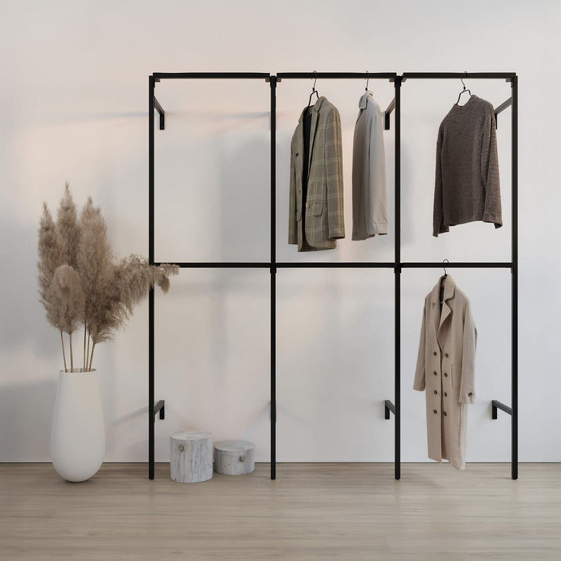 Schwarze Kleiderstange mit vier Kleidungsstücken in neutralen Farben vor weißer Wand und Vase mit Pampasgras auf hellem Boden