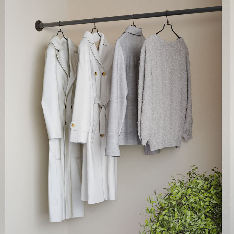 Vier helle Kleidungsstücke, darunter Mäntel und Pullover, an einer dunklen Stange vor hellgrauer Wand mit Pflanze unten rechts