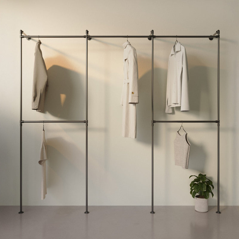 Schwarzes Kleidergestell mit hellen Kleidungsstücken und grünem Topf mit Pflanze vor beige Wand in minimalistischem Raum