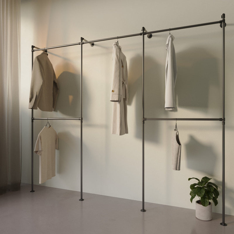 Minimalistische Kleiderstange aus Metall mit wenigen hellen Kleidungsstücken vor beigeweißer Wand und Pflanzenübertopf